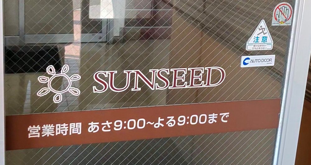 サンシード食品館の入口のドア。「SUNSEED」の文字と「営業時間 あさ9:00〜よる9:00まで」