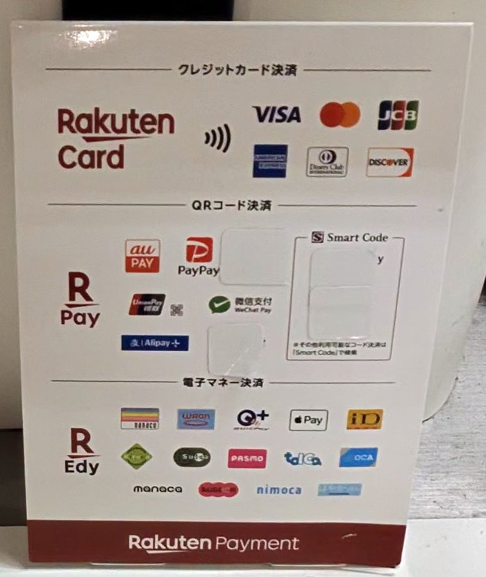 レジに置かれた楽天ペイメントの支払い方法の案内。クレジットカード・QRコード決済・電子マネーのマークが並び、一部のマークは白いシールで隠されている