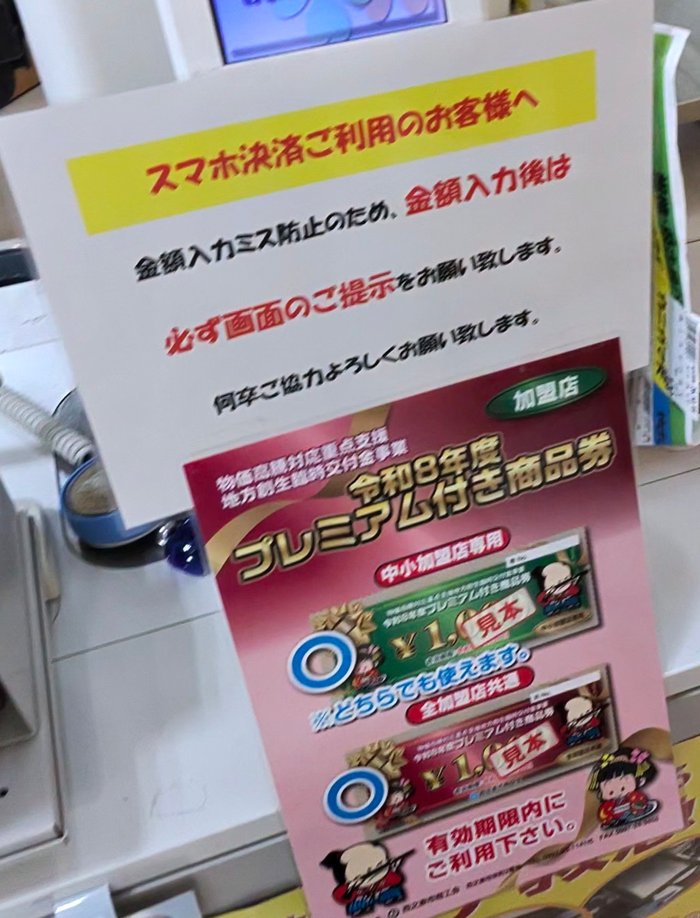 セルフレジに貼られた「スマホ決済ご利用のお客様へ 金額入力後は必ず画面のご提示を」の案内と、西之表市 令和8年度プレミアム付き商品券の加盟店の掲示
